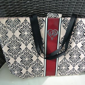 May include: A patterned tote bag with a cream background and black floral design. The bag features black straps and a central red stripe with a silver heart emblem. The bag is on a white surface.