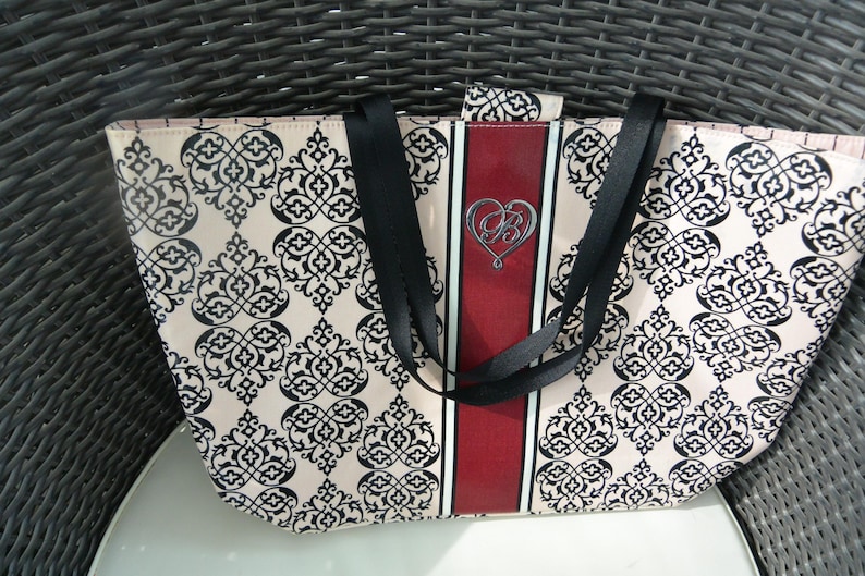 May include: A patterned tote bag with a cream background and black floral design. The bag features black straps and a central red stripe with a silver heart emblem. The bag is on a white surface.