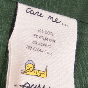 May include: A white care label with black text that reads "care me... 60% wool 18% poliamide 22% acrylic dry clean only" and a cartoon cat illustration. The label is attached to a dark green fabric.