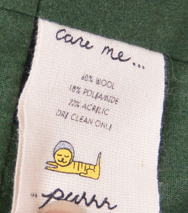 May include: A white care label with black text that reads "care me... 60% wool 18% poliamide 22% acrylic dry clean only" and a cartoon cat illustration. The label is attached to a dark green fabric.