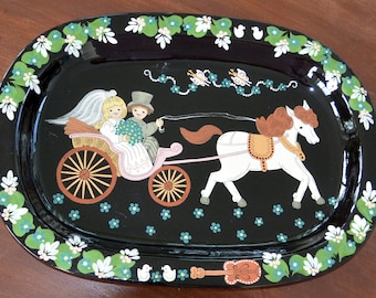 May include: A black oval serving tray featuring a painted scene of a bride and groom riding in a horse-drawn carriage. The carriage is pink with white wheels and the horse is white with a brown mane and tail. The tray is decorated with green leaves, white flowers, and blue forget-me-nots.