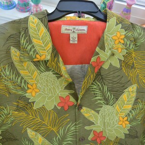 K&ouml;nnte beinhalten: Olivgr&uuml;nes Hemd mit tropischem Blumenmuster. Das Hemd hat gro&szlig;e gr&uuml;ne Bl&auml;tter mit gelben Akzenten, orangefarbene und gelbe Bl&uuml;ten und ein rotes Innenfutter. Das Hemd h&auml;ngt an einem schwarzen Kleiderb&uuml;gel.