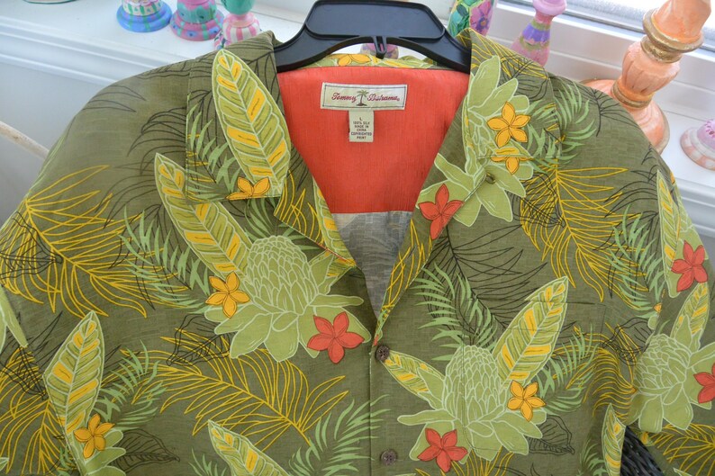 K&ouml;nnte beinhalten: Olivgr&uuml;nes Hemd mit tropischem Blumenmuster. Das Hemd hat gro&szlig;e gr&uuml;ne Bl&auml;tter mit gelben Akzenten, orangefarbene und gelbe Bl&uuml;ten und ein rotes Innenfutter. Das Hemd h&auml;ngt an einem schwarzen Kleiderb&uuml;gel.