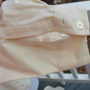 Puede incluir: Primer plano del pu&ntilde;o de una camisa amarillo claro. El pu&ntilde;o tiene cierre de bot&oacute;n, ojal y una tira de tela. La tela tiene una textura sutil.