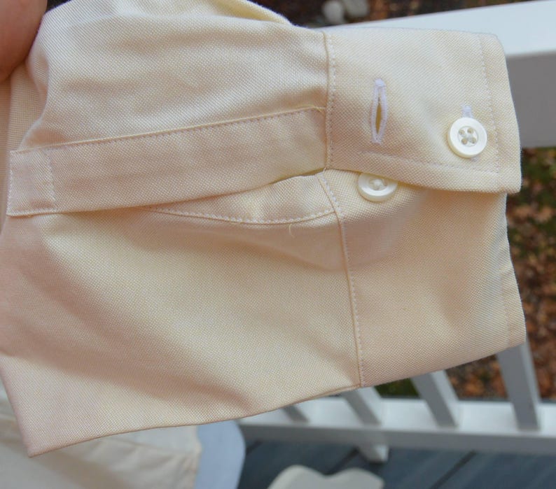 Puede incluir: Primer plano del pu&ntilde;o de una camisa amarillo claro. El pu&ntilde;o tiene cierre de bot&oacute;n, ojal y una tira de tela. La tela tiene una textura sutil.