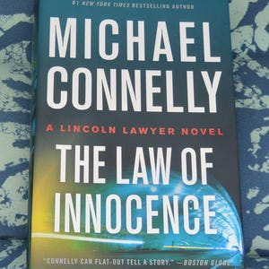 May include: A hardcover book titled "The Law of Innocence" by Michael Connelly. The cover is predominantly dark blue with white lettering. The book is a "Lincoln Lawyer Novel" and is a #1 New York Times Bestselling Author.