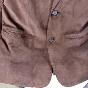 May include: A brown suede blazer with two buttons and two front pockets. The blazer is a solid color and appears to be made of a soft, textured material. The blazer is resting on a wicker chair.