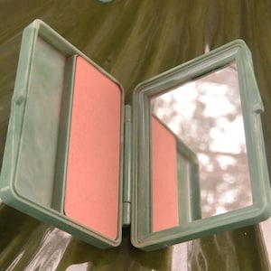 May include: Open, vintage-style compact in a light green hue. The compact is divided into two sections, one with a peach-colored powder and the other with a mirror. The compact is open, revealing its contents.