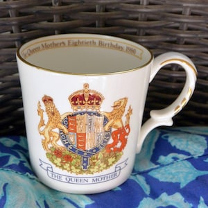 May include: White ceramic mug with gold trim and a handle. The mug features a royal crest with lions, a crown, and the text "The Queen Mother" and "The Queen Mother's Eightieth Birthday, 1980".