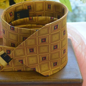 Puede incluir: Una corbata dorada enrollada con un patrón geométrico repetitivo de cuadrados en tonos dorados, rojos y azul marino. La etiqueta dice "OTT". La corbata está sobre una superficie de madera.