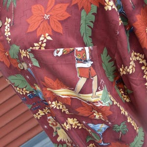 May include: A maroon button-up shirt with a festive Hawaiian print. The design features red poinsettias, green leaves, and illustrations of surfers, cocktails, and tropical scenes. The shirt has a pocket with a surfer graphic.