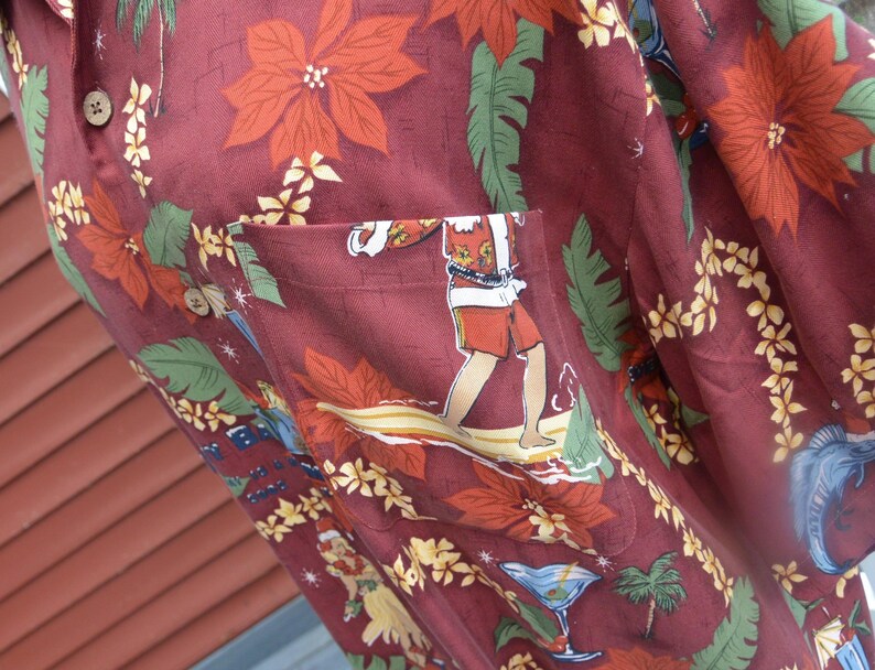 May include: A maroon button-up shirt with a festive Hawaiian print. The design features red poinsettias, green leaves, and illustrations of surfers, cocktails, and tropical scenes. The shirt has a pocket with a surfer graphic.