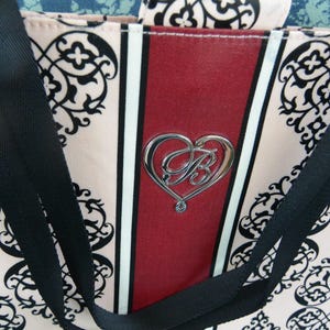 May include: A tote bag featuring a pink and black floral design. The bag has a central red stripe with a silver heart-shaped emblem. Black straps are attached to the top of the bag.