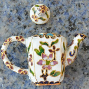 May include: A small, white decorative teapot with a matching lid. The teapot is adorned with floral designs in pink, green, and purple, outlined in gold. The handle and spout are also detailed with gold accents.