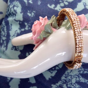 May include: A gold bangle bracelet with a row of white pearl-like beads. The bracelet is on a white porcelain hand with a pink flower.