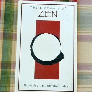 May include: A book titled "The Elements of Zen" with a white cover, a red rectangle, and a black circle design. The authors' names, David Scott & Tony Doubleday, are printed at the bottom. The book is on a colorful plaid surface.