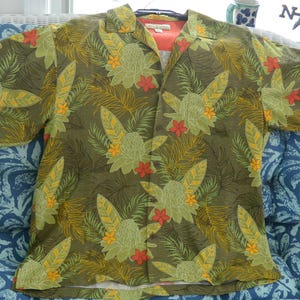 K&ouml;nnte beinhalten: Ein kurz&auml;rmeliges Hemd mit tropischem Blumenmuster. Das Hemd hat einen gr&uuml;nen Hintergrund mit gro&szlig;en gr&uuml;nen, gelben und roten Blumen- und Blattmustern. Das Hemd ist offen und zeigt ein pfirsichfarbenes Unterhemd.