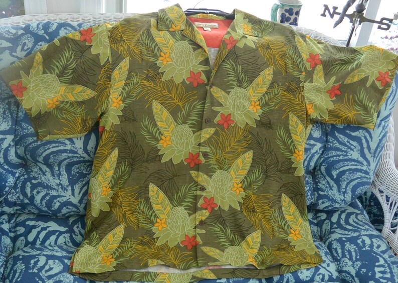 K&ouml;nnte beinhalten: Ein kurz&auml;rmeliges Hemd mit tropischem Blumenmuster. Das Hemd hat einen gr&uuml;nen Hintergrund mit gro&szlig;en gr&uuml;nen, gelben und roten Blumen- und Blattmustern. Das Hemd ist offen und zeigt ein pfirsichfarbenes Unterhemd.