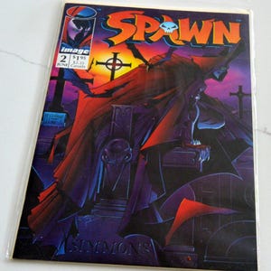 Op de afbeelding: Een Spawn stripboek nummer 2, uitgegeven door Image Comics in juni. De cover toont een donker figuur met een cape op een begraafplaats met een zonsondergang, de titel Spawn in oranje en de naam Simmons onderaan de pagina.