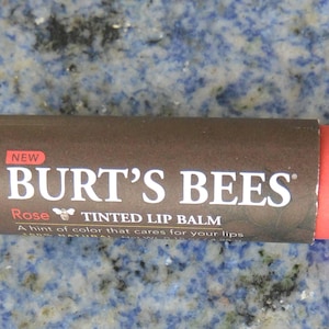 May include: A brown tube of Burt's Bees Rose Tinted Lip Balm. The tube has a red cap and the text "NEW" is printed on the tube.