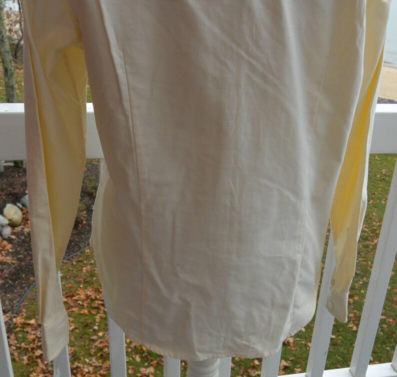 Puede incluir: Camisa de manga larga de color amarillo p&aacute;lido con pu&ntilde;o abotonado. La camisa est&aacute; hecha de una tela suave y tiene un corte entallado. La parte trasera de la camisa es visible, mostrando las costuras y la forma de la prenda.