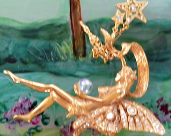May include: A gold-toned fairy figurine with a star-shaped crown and wings adorned with rhinestones. The fairy is holding a blue iridescent gem and is flying with her legs outstretched.