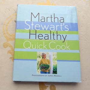 May include: A cookbook titled "Martha Stewart's Healthy Quick Cook" with a light blue cover and horizontal stripes of blue and green. The cover features a photo of Martha Stewart holding a basket of produce. The book's title is in large, dark brown letters.