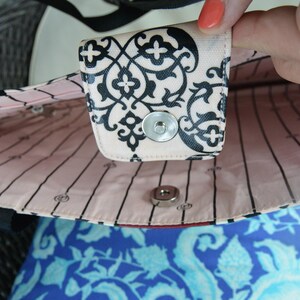 May include: Close-up of a tote bag interior. The bag has a light pink lining with black vertical stripes and a decorative black and white patterned flap with a snap closure. The bag's exterior is black.