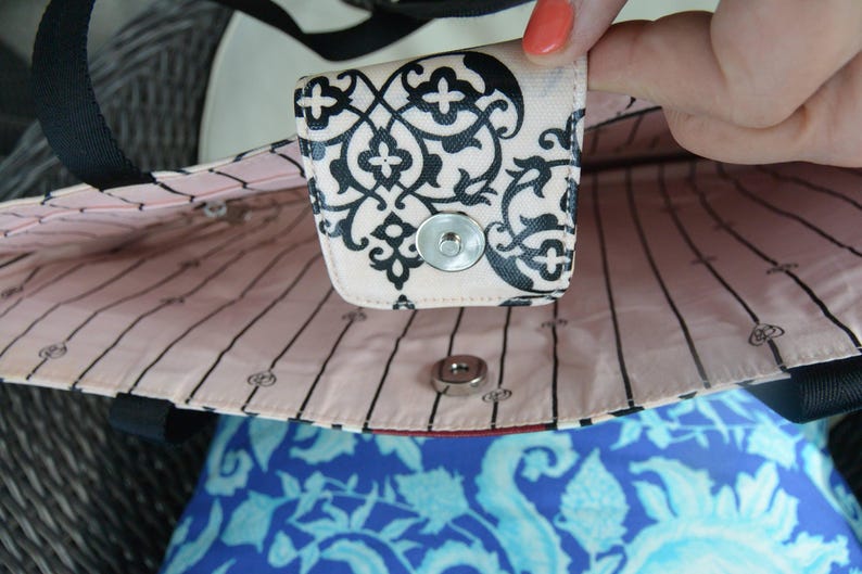 May include: Close-up of a tote bag interior. The bag has a light pink lining with black vertical stripes and a decorative black and white patterned flap with a snap closure. The bag's exterior is black.