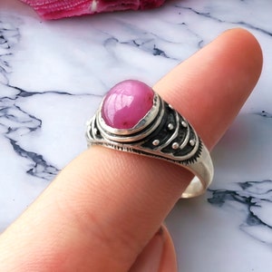 May include: A silver ring with a pink gemstone set in a decorative bezel. The ring has a textured band with small silver beads.