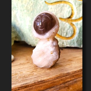 May include: A small, carved crystal figurine with a human-like face. The figurine is made of a light pink crystal with a dark brown face. The figurine is standing on a wooden surface.