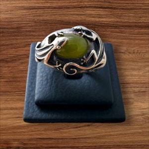 May include: A silver ring with a green gemstone set in a detailed design of a lizard. The ring is displayed on a black ring stand.