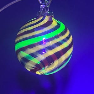 May include: A round, glass ornament with a striped pattern of yellow, maroon, and green. The ornament is suspended by a clear glass loop and wire against a dark blue background. The glass has a reflective quality.
