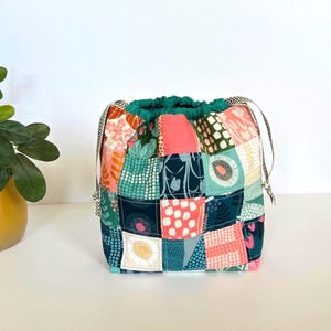 May include: A drawstring pouch constructed from a patchwork of quilted fabric squares featuring various patterns in shades of teal, coral, pink, and white. The bag has a solid teal fabric top edge and two patterned fabric drawstrings on each side.