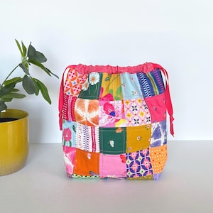 May include: A patchwork drawstring bag with a vibrant mix of floral and geometric patterns in pink, yellow, green, and blue. The bag has a pink drawstring closure and red ties. The bag is sitting on a white surface.