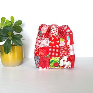 May include: A patchwork drawstring bag in various shades of red, featuring floral, polka dot, and geometric patterns. The bag is made of quilted fabric and sits on a white surface. A yellow potted plant is in the background.