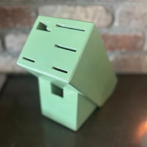 May include: A light green wooden knife block with multiple slots for knife storage. The block is rectangular with a slightly angled top and a square base. The background features a brick wall.