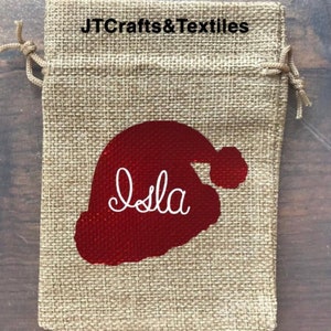 May include: A burlap gift bag with a red Santa hat design and the name "Isla" embroidered in white. The bag has a drawstring closure.