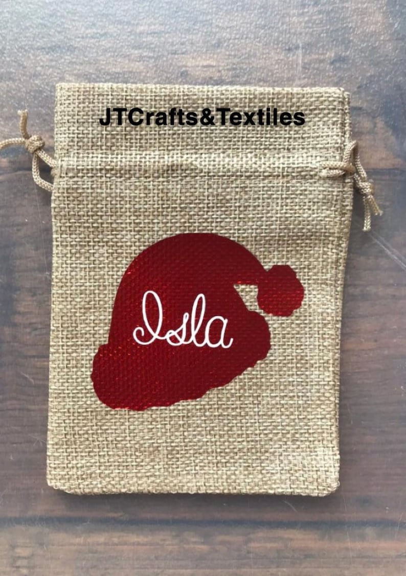 May include: A burlap gift bag with a red Santa hat design and the name "Isla" embroidered in white. The bag has a drawstring closure.