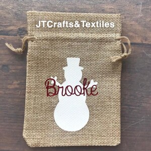 May include: A burlap gift bag with a white snowman silhouette and the name "Brooke" in red glitter lettering. The bag has a drawstring closure and is perfect for small gifts.