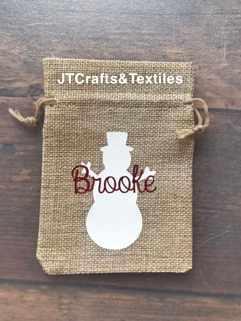 May include: A burlap gift bag with a white snowman silhouette and the name "Brooke" in red glitter lettering. The bag has a drawstring closure and is perfect for small gifts.