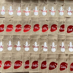 May include: A collection of 40 burlap gift bags with personalized names in red and white. The bags feature a snowman or Santa hat design.