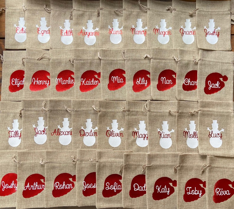 May include: A collection of 40 burlap gift bags with personalized names in red and white. The bags feature a snowman or Santa hat design.