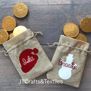 May include: Two small burlap gift bags with personalized names, "Isla" and "Brooke", and festive holiday designs. The bag with "Isla" has a red Santa hat design, and the bag with "Brooke" has a white snowman design.  Each bag is filled with gold chocolate coins.