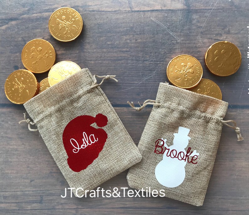 May include: Two small burlap gift bags with personalized names, "Isla" and "Brooke", and festive holiday designs. The bag with "Isla" has a red Santa hat design, and the bag with "Brooke" has a white snowman design.  Each bag is filled with gold chocolate coins.