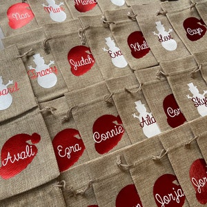 May include: A collection of burlap gift bags with red and white snowman and Santa Claus designs. The bags are personalized with names such as Marco, Mana, Ramirez, Agina, Finn, Nan, Mur, Judah, Eva, Khal, Levi, Cayo, Avali, Ezra, Connie, Aild, Cor, Sky, Seth, Conn, Jorja, and Jor.