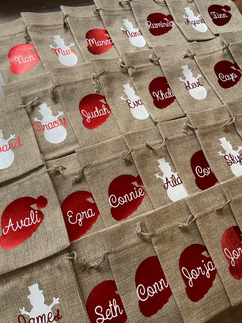 May include: A collection of burlap gift bags with red and white snowman and Santa Claus designs. The bags are personalized with names such as Marco, Mana, Ramirez, Agina, Finn, Nan, Mur, Judah, Eva, Khal, Levi, Cayo, Avali, Ezra, Connie, Aild, Cor, Sky, Seth, Conn, Jorja, and Jor.