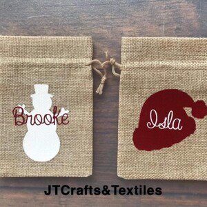 May include: Two burlap gift bags with personalized Christmas designs. One bag features a white snowman with the name "Brooke" in red lettering. The other bag features a red Santa hat with the name "Isla" in white lettering. The bags are on a wooden surface.