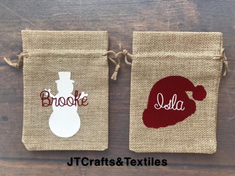 May include: Two burlap gift bags with personalized Christmas designs. One bag features a white snowman with the name "Brooke" in red lettering. The other bag features a red Santa hat with the name "Isla" in white lettering. The bags are on a wooden surface.