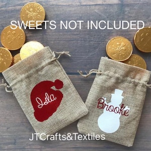 May include: Two small burlap gift bags with personalized Christmas designs. The bag on the left has a red Santa hat with the name "Isla" written in white. The bag on the right has a white snowman with the name "Brooke" written in red. The text "SWEETS NOT INCLUDED" is visible at the top of the image.